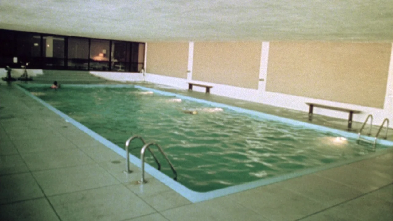 Shivers (1975) movie still: wide — An indoor rectangular swimming pool features pale tiled decks and dark wall-to-ceiling windows. A lone swimmer…