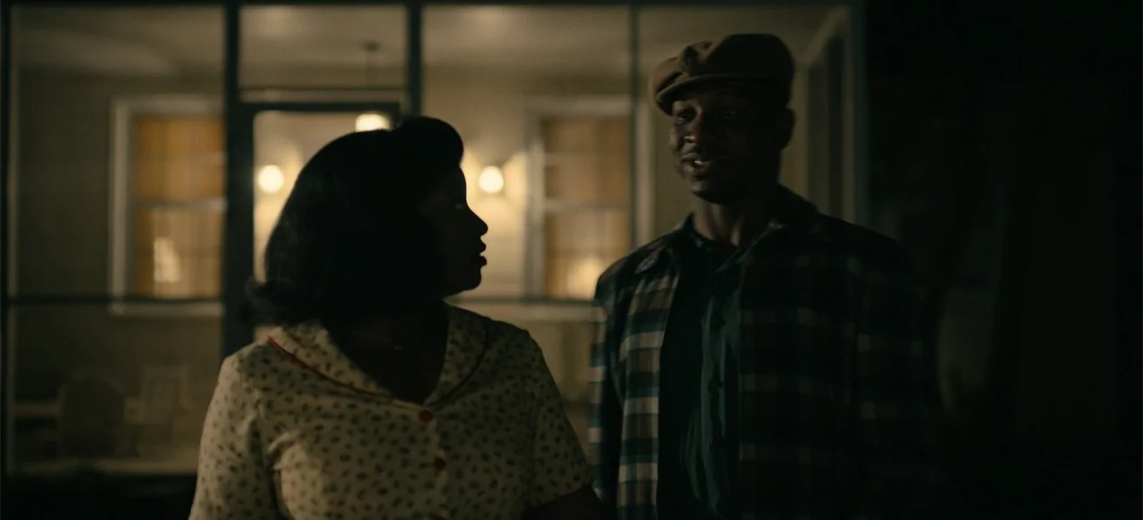 Devotion (2022) movie still: medium — A dark house porch at night with illuminated windows in the background. A woman and a man face each other…