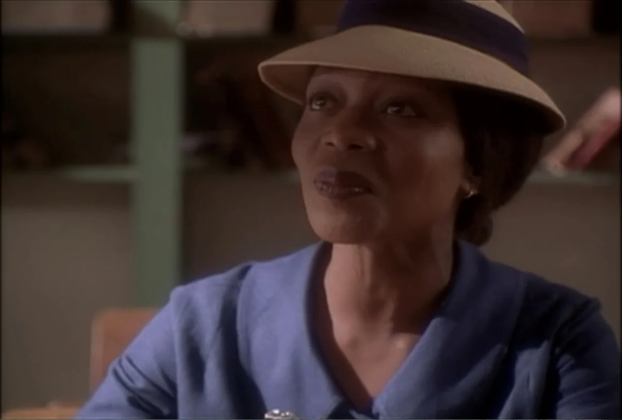 Miss Evers' Boys (1997) movie still: medium — An interior space features blurred shelves with books in the background. A woman in a blue dress and…