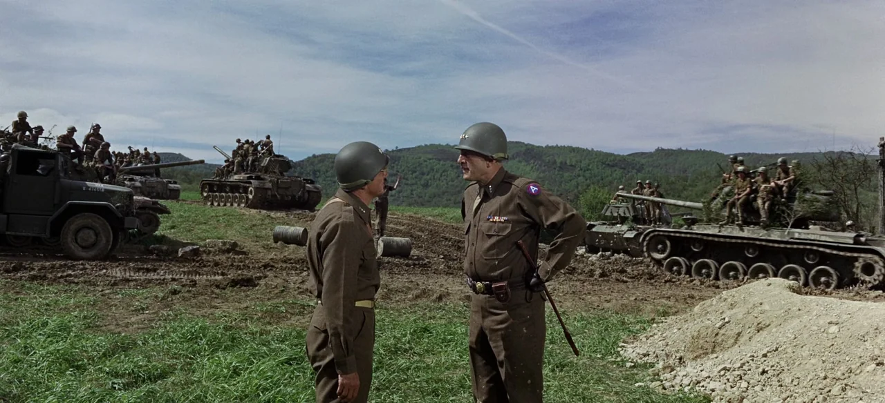 Patton (1970) movie still: fullbody — Two military officers in olive drab uniforms stand in a grassy muddy field. Several tanks and soldiers are…