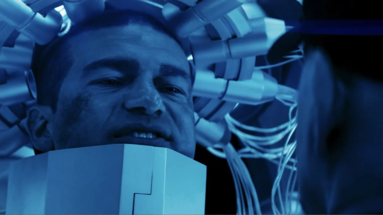 Robot Overlords (2015) movie still: closeup — A dark, technological lab space features a man fitted with a complex cranial device. He lies in a…