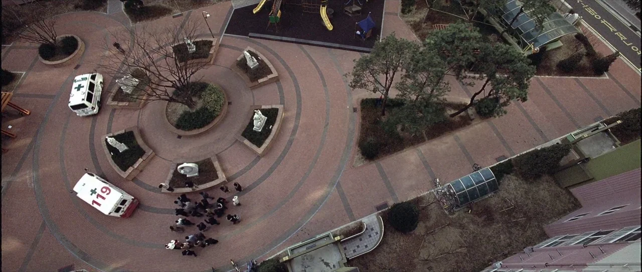 Lady Vengeance (2005) movie still: establishing — A top-down view of a circular brick-paved public plaza featuring white stone sculptures. Two…
