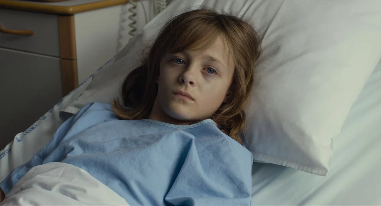 Happy End (2017) movie still: closeup — A sterile medical room features a white pillow and a wooden bedside cabinet. A young child wearing a blue…