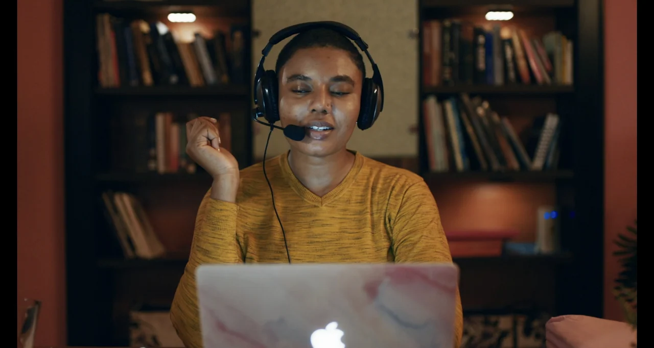 Love In A Pandemic (2023) movie still: medium — A person sits in a home office with a dark bookshelf filled with books. The subject wears a headset…