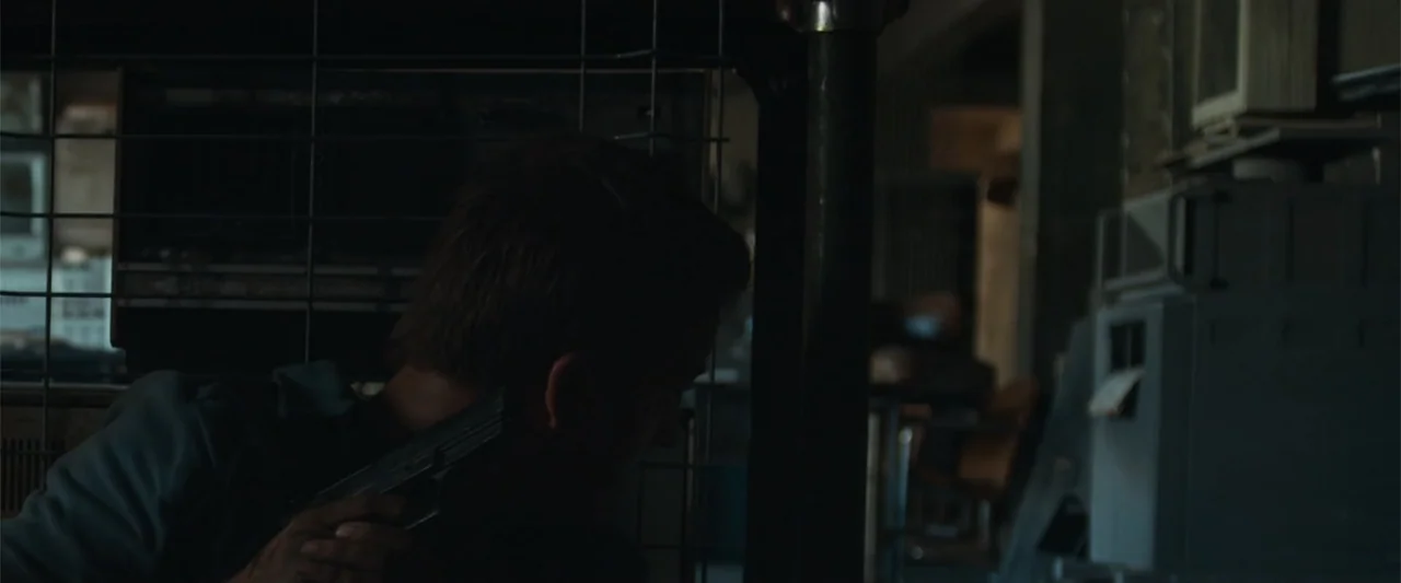 The Condemned 2 (2015) movie still: medium — A dimly lit industrial office space features a metal mesh partition in the foreground. A man in a dark…