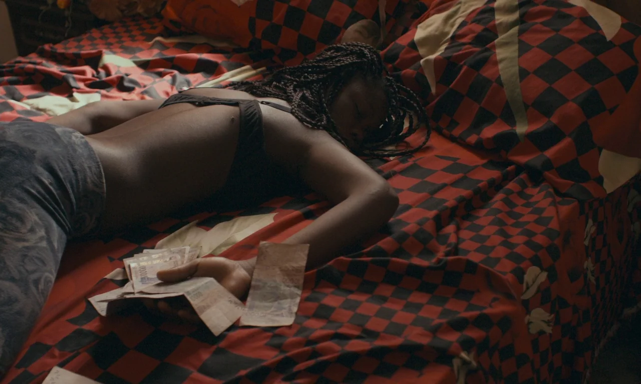 Atlantics (2019) movie still: medium — A person lies across a bed featuring a red and black checkered bedspread. They hold a small stack of paper…