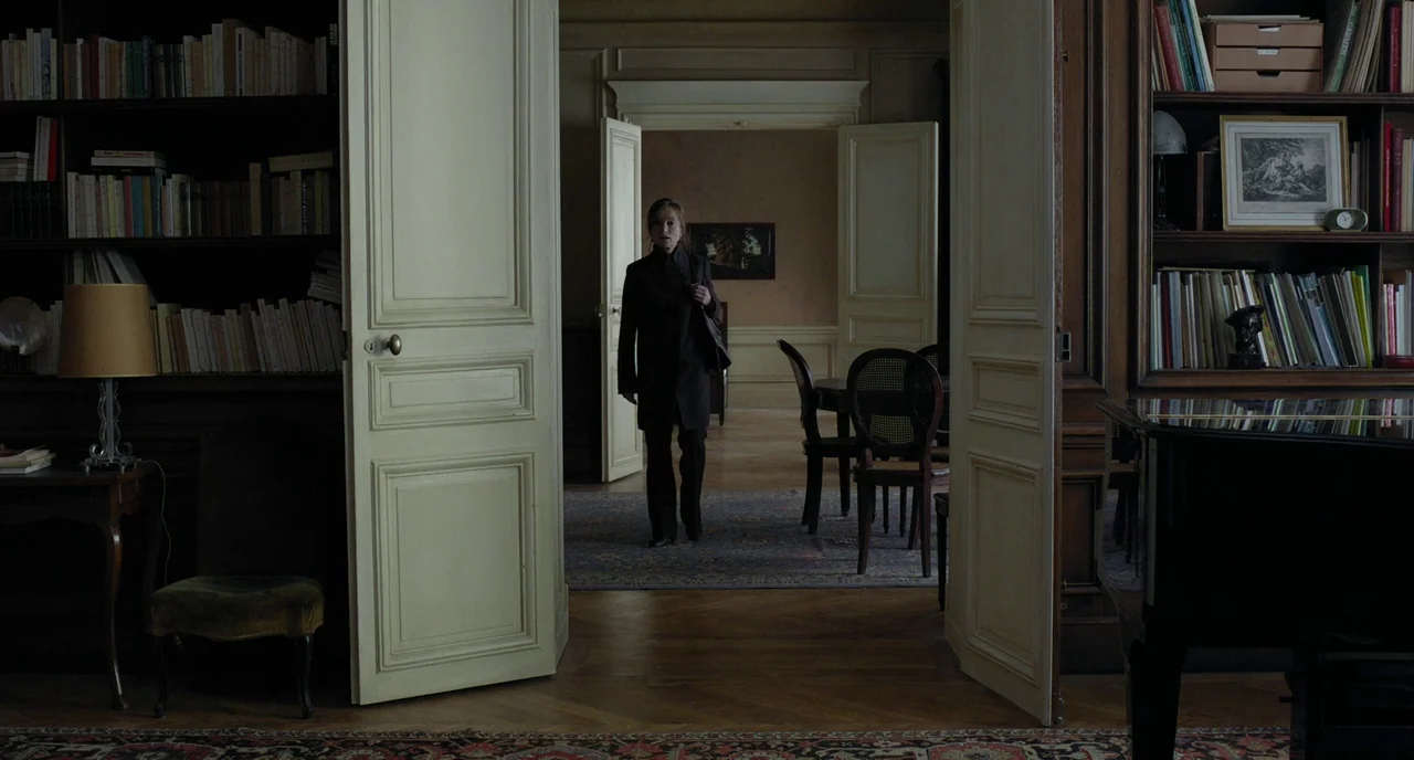 Amour (2012) movie still: wide — A grand, multi-room interior with tall doors, built-in bookcases, and wood flooring. A woman in dark clothing walks…