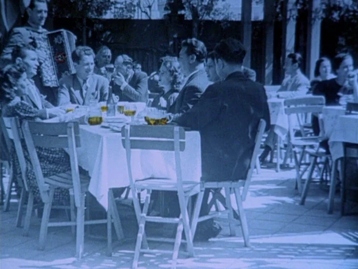 Innocence Unprotected (1968) movie still: medium — An outdoor seating area with white-clothed tables sits on a paved patio under sunlight. A man…