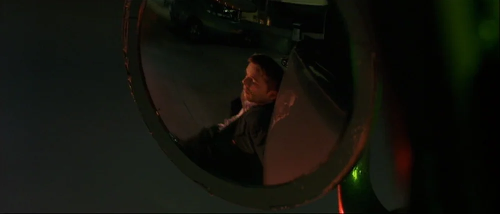 Gattaca (1997) movie still: closeup — A man is reflected in the circular side mirror of a dark vehicle at night. He is slumped against the car frame…