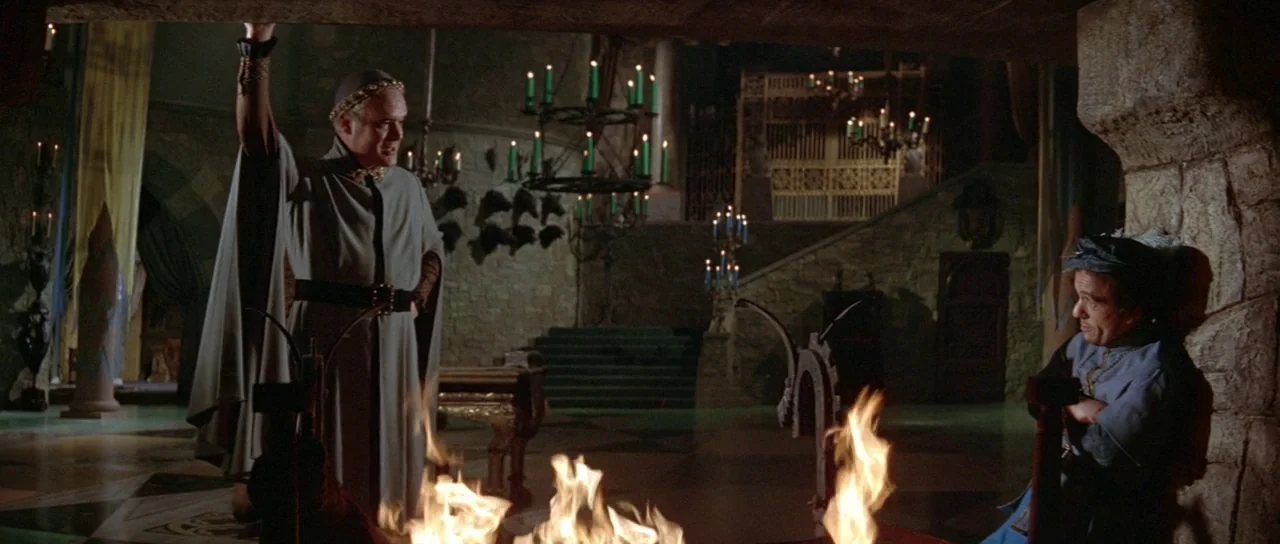 The Masque of the Red Death (1964) movie still: wide — A dark stone hall features ornate iron chandeliers holding green candles and high ceilings. A…