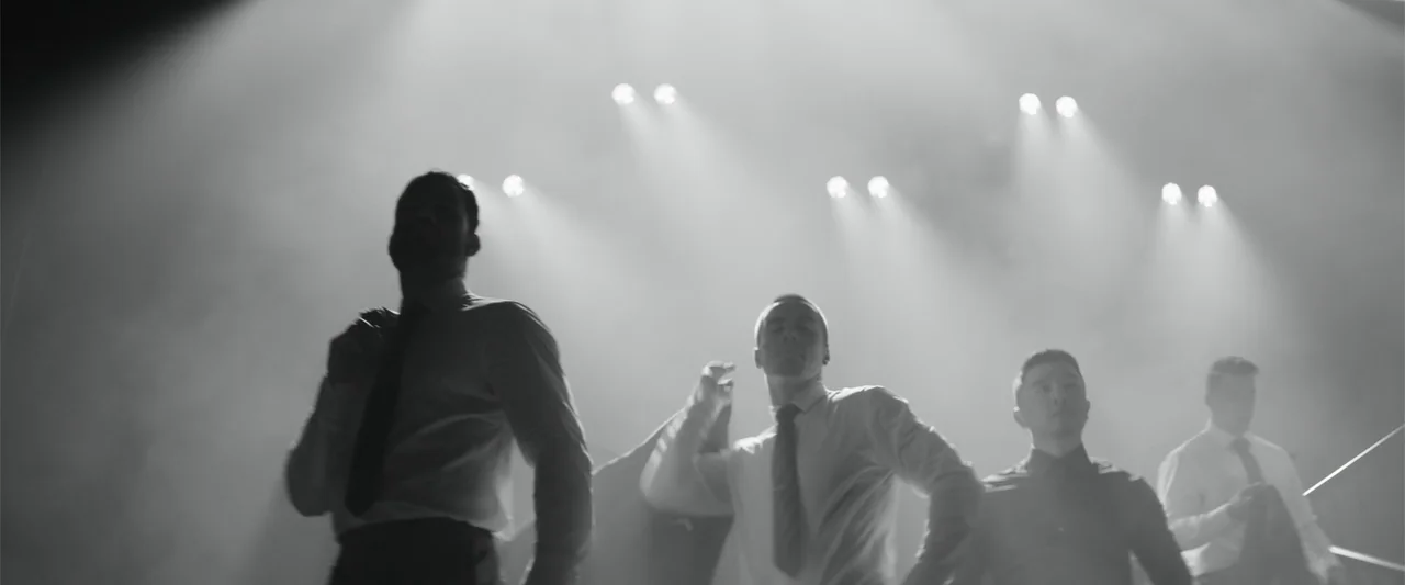 Magic Mike's Last Dance (2023) movie still: medium — A dimly lit stage filled with haze and sharp overhead spotlights. Four men in suits move in…