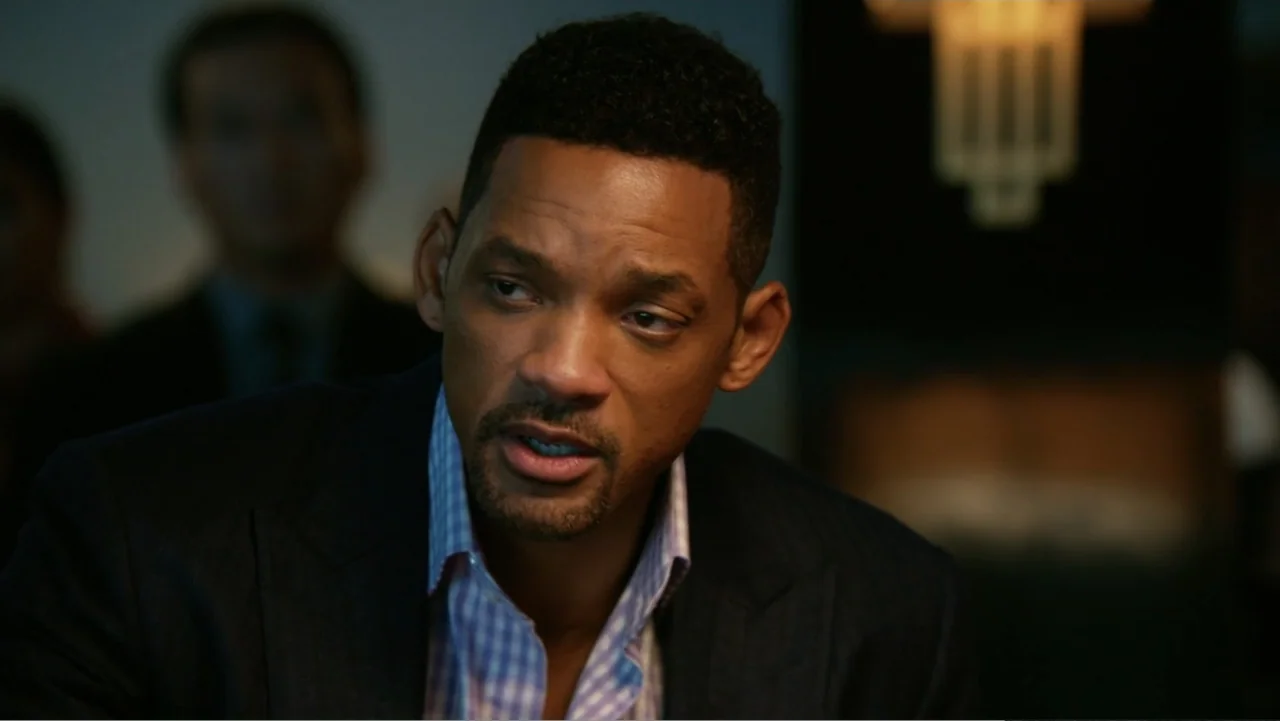 Focus (2015) movie still: closeup — An indoor office environment with a dark background and a blurred light fixture. A man in a blazer and checkered…