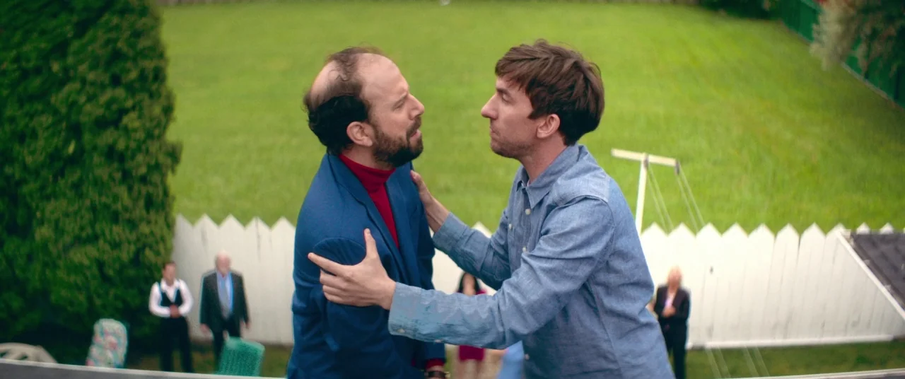Room For Rent (2017) movie still: medium — A suburban backyard features a white picket fence and a bright green lawn. Two men face each other in an…