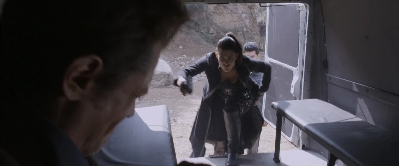 Skiptrace (2016) movie still: medium — A woman in a black coat steps from a van into a rocky area. She holds a handgun while emerging from the…