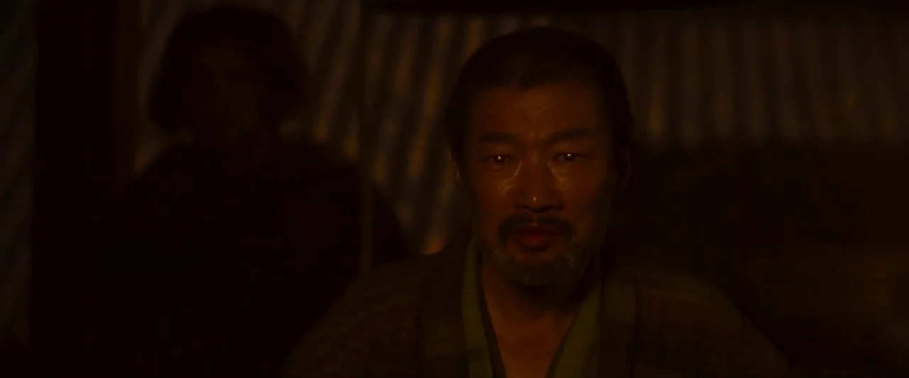 Kubi (2023) movie still: closeup — A dimly lit interior space with vertical fabric panels visible in the background. An middle-aged man with a…