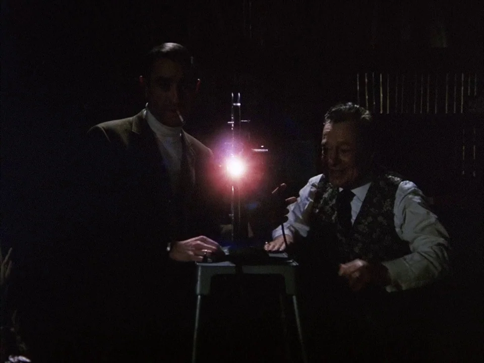 The Clowns (1970) movie still: medium — A dark, dimly lit room contains two men operating a vintage movie projector. The bright light from the…