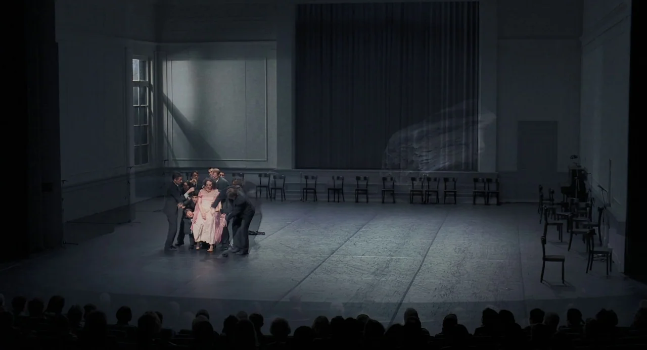Pina (2011) movie still: wide — A minimalist auditorium stage contains a group of formally dressed figures near the left side. A woman in a pink…