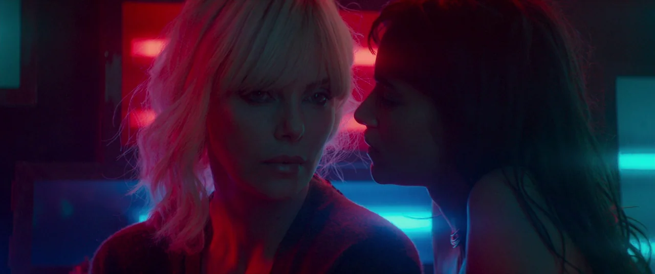Atomic Blonde (2017) movie still: closeup — A dark space lit by sharp red and blue neon light sources. Two women face each other in close proximity…