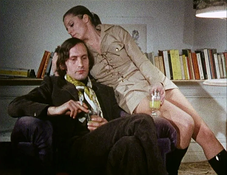 Rio Das Mortes (1971) movie still: medium — A 1970s interior with bookshelves lining the wall behind a plush upholstered chair. A man sits in the…