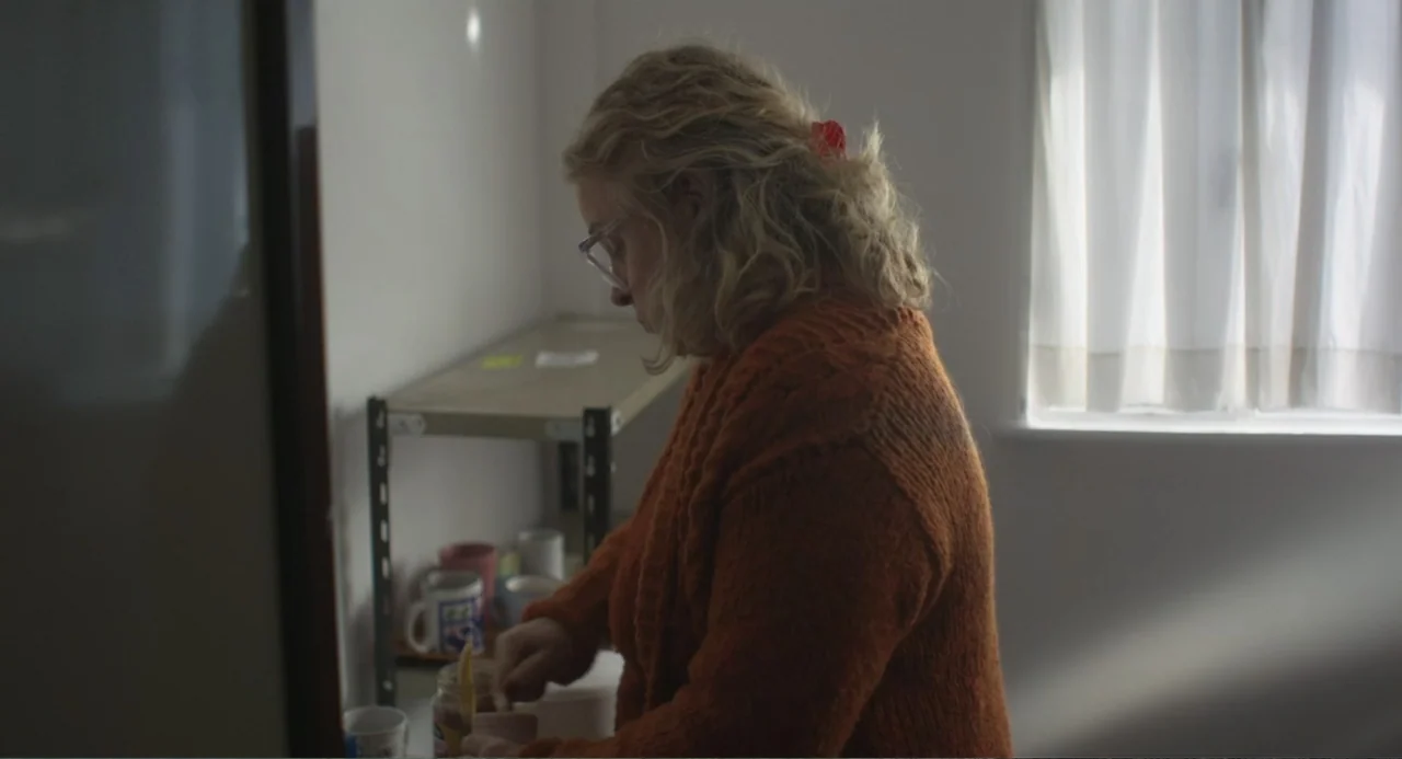 An Odd Turn (2024) movie still: medium — A spare kitchen features a simple metal shelving unit holding various ceramic mugs. A woman in a textured…