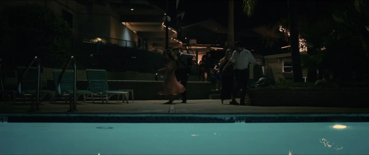 Plus One (2019) movie still: wide — An exterior poolside area at night with lounge chairs and terraced building architecture visible. A small group…