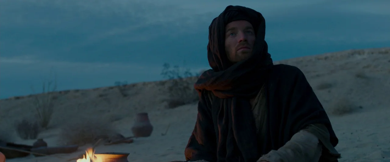 Last Days in the Desert (2015) movie still: medium — A man wearing a hooded cloak sits in a dark, sprawling sandy desert at night. A small campfire…