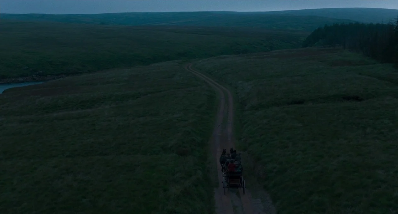 Peterloo (2018) movie still: wide — A dark, hilly moorland landscape stretches under a dim blue-hour sky. A small horse-drawn carriage travels…
