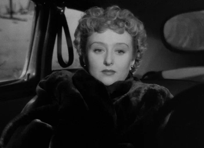 All About Eve (1950) movie still: closeup — The interior of a vintage automobile at night with a dark backdrop. A woman with curled hair sits…