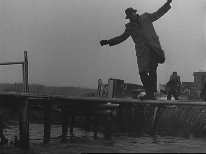 Blast Of Silence (1961) movie still: fullbody — A wooden plank stretches over a narrow waterway in a marshy environment. A man carefully walks…