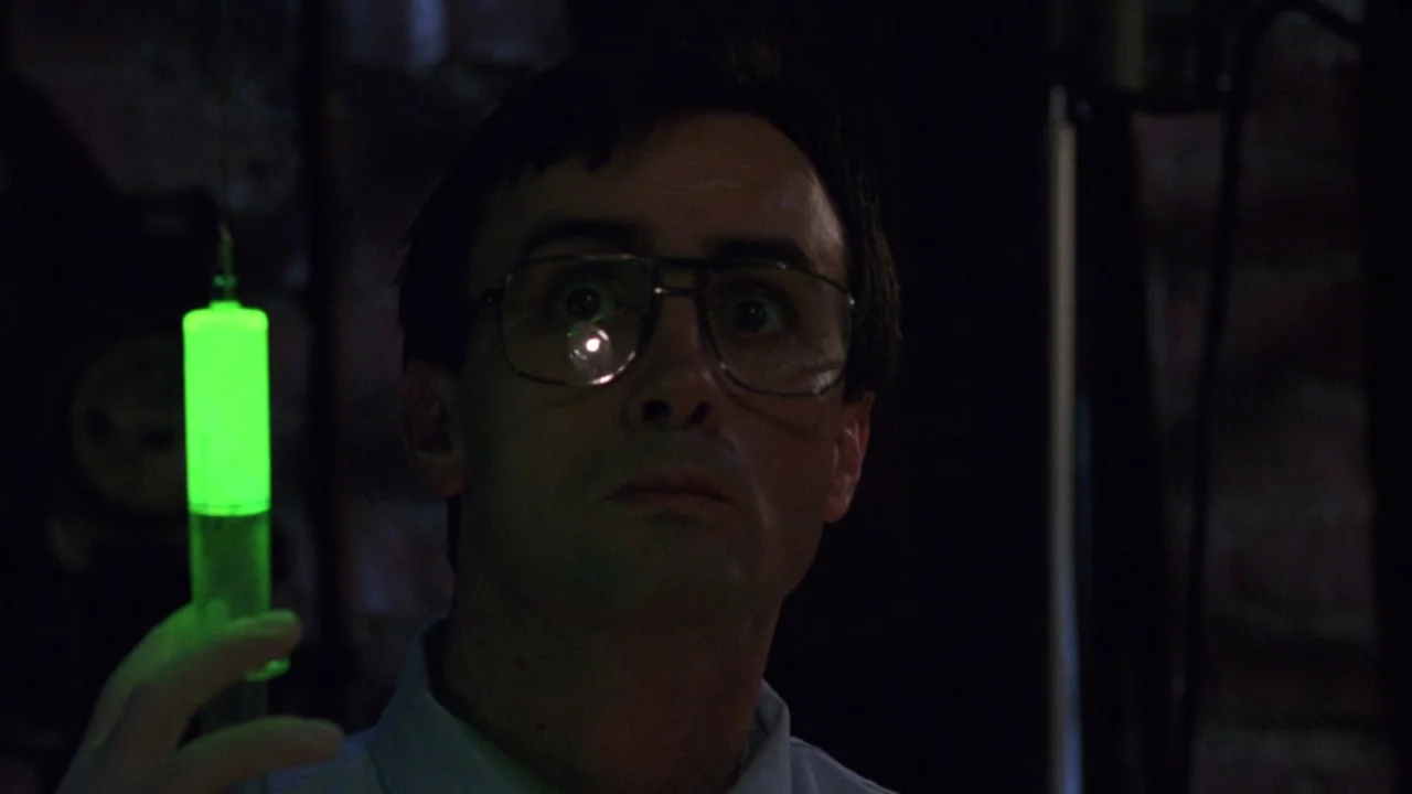 Bride of Reanimator (1989) movie still: closeup — A dimly lit room with dark surroundings and exposed brick textures. A man in glasses observes a…