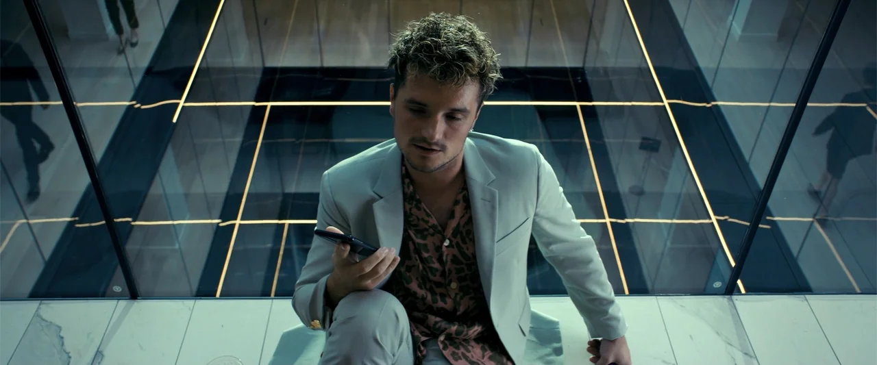 The Beekeeper (2024) movie still: medium — A man sits on a marble ledge within a modern minimalist atrium space. He looks down at his smartphone…
