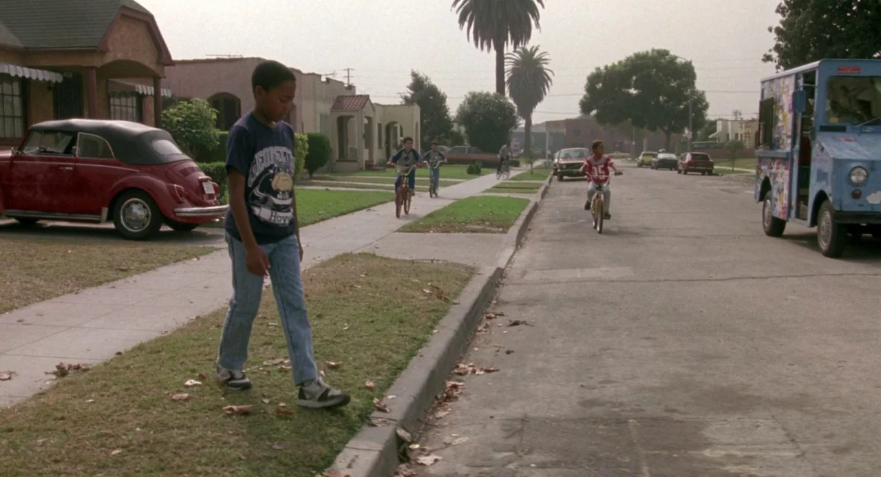 Boyz In The Hood (1991) movie still: wide — A residential street in a low-density suburban neighborhood with parked cars and sidewalks. A young boy…