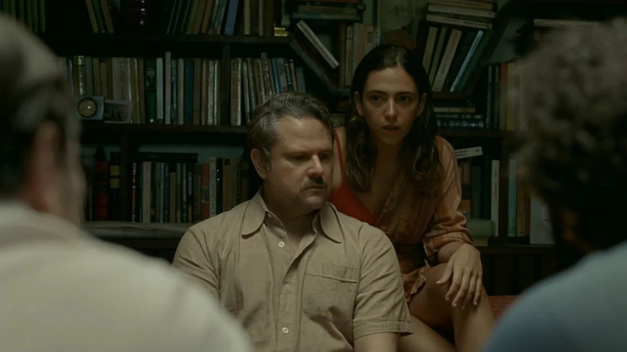 I'm Still Here (2024) movie still: medium — A home library featuring dark wood shelves densely packed with various colorful books. A man and a woman…