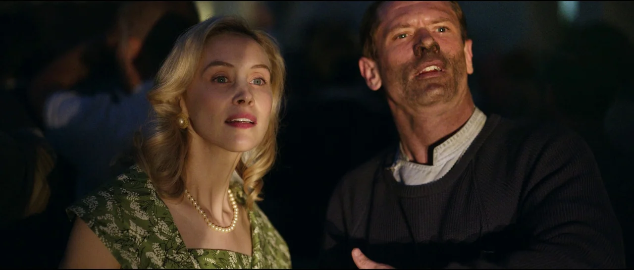 Ferrari (2023) movie still: medium — A dark outdoor setting at night features two people illuminated by low-angle light. A woman in a floral dress…