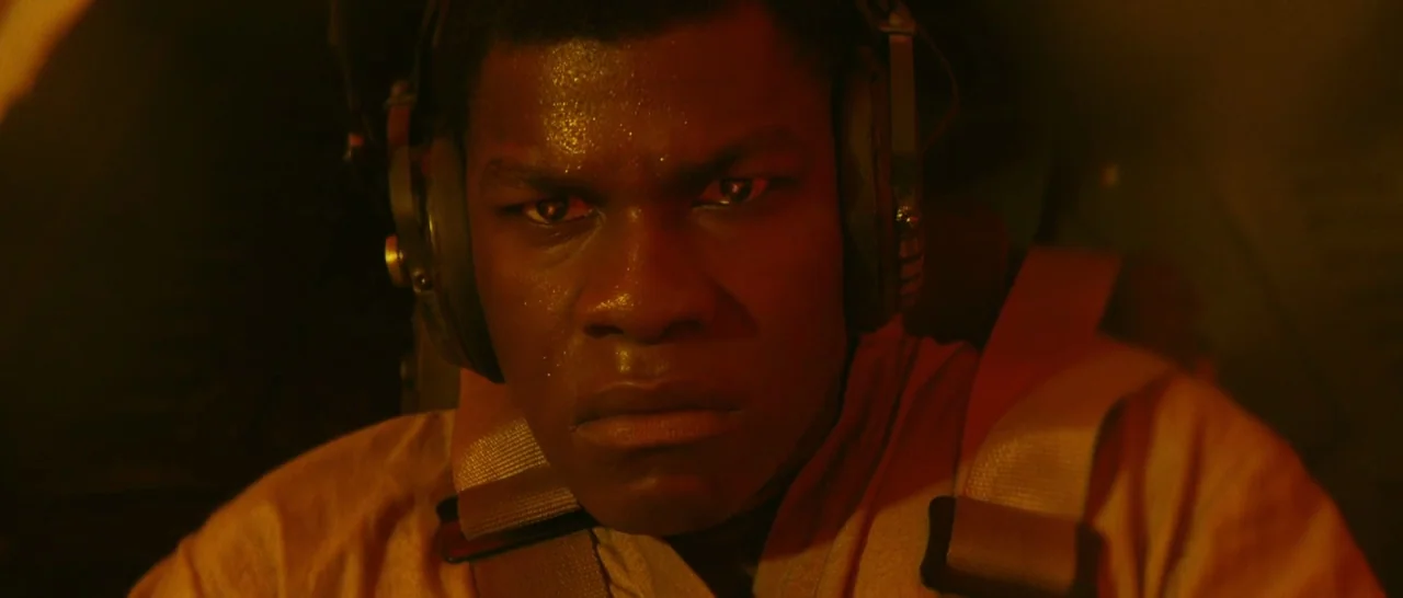 Star Wars: The Last Jedi (2017) movie still: closeup — A dimly lit interior cockpit bathed in warm amber emergency illumination. A young man wearing…