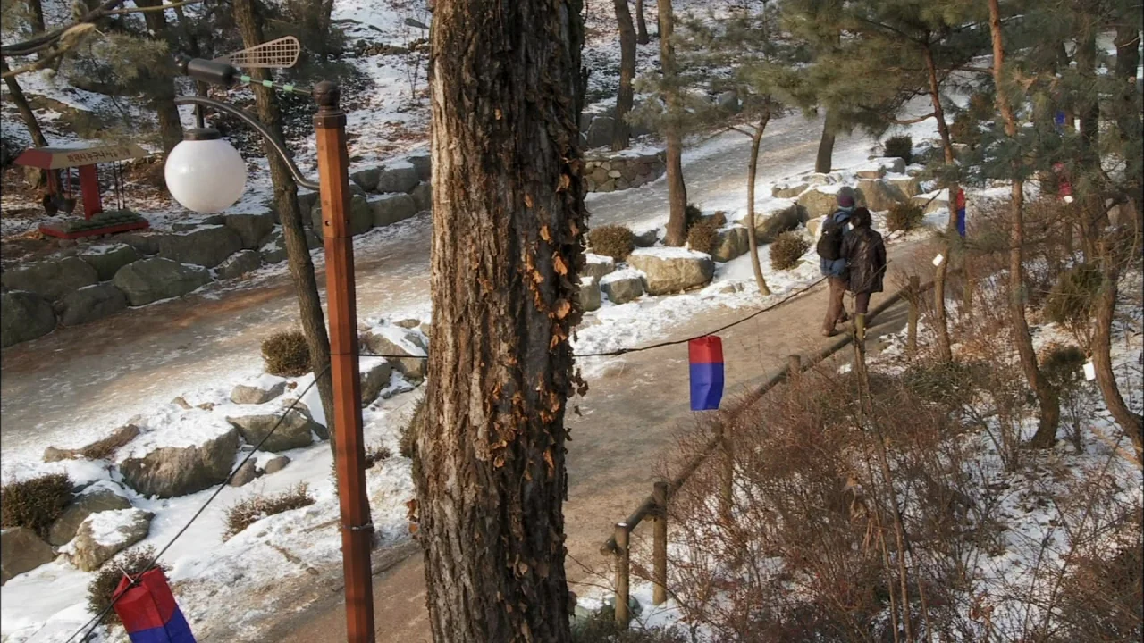 Oki’s Movie (2010) movie still: wide — A snow-dusted park path winds between bare trees and rocky terrain in winter. Two people walk away from the…