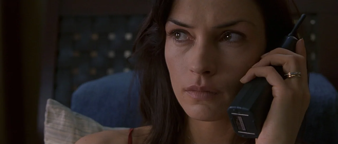 Don't Say a Word (2001) movie still: closeup — A woman is indoors within a dimly lit space featuring a dark headboard. She holds a black cordless…