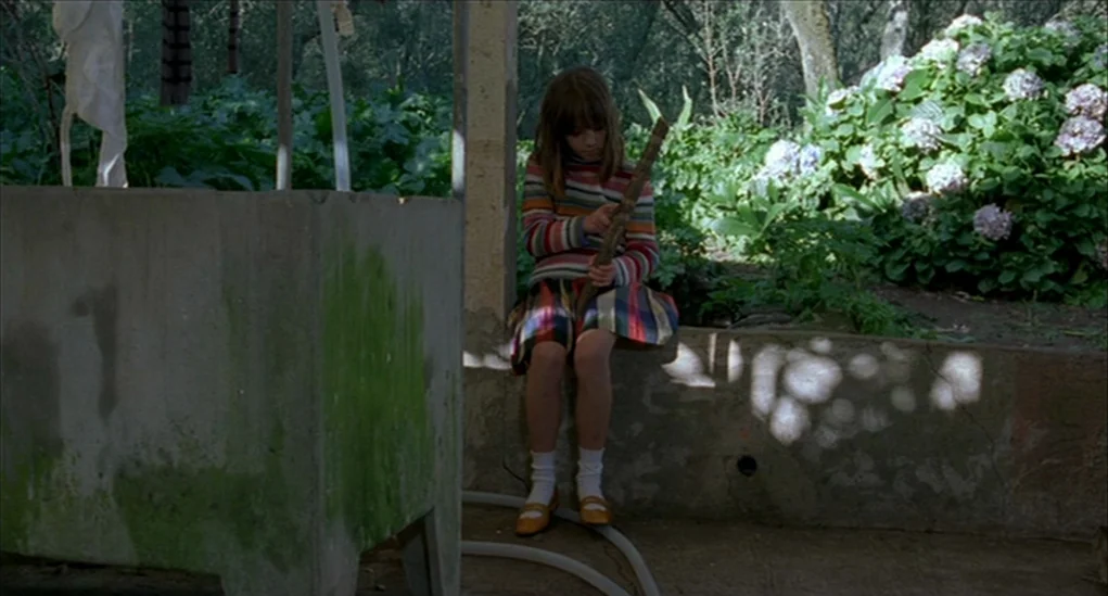 Anatomy of Hell (2004) movie still: fullbody — An outdoor area features a mossy concrete basin beside a low stone wall. A young girl in a striped…