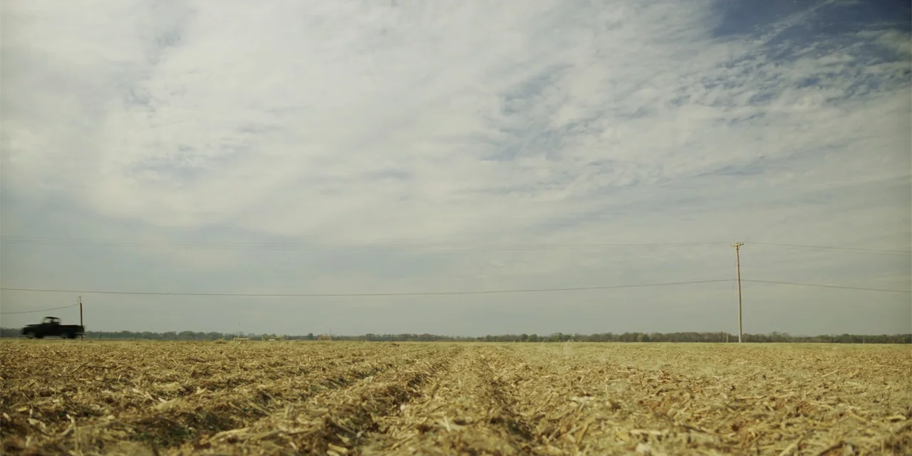 Green Book (2018) movie still: establishing — A vast, harvested agricultural field stretches toward the horizon under a wide, cloudy sky. A small…