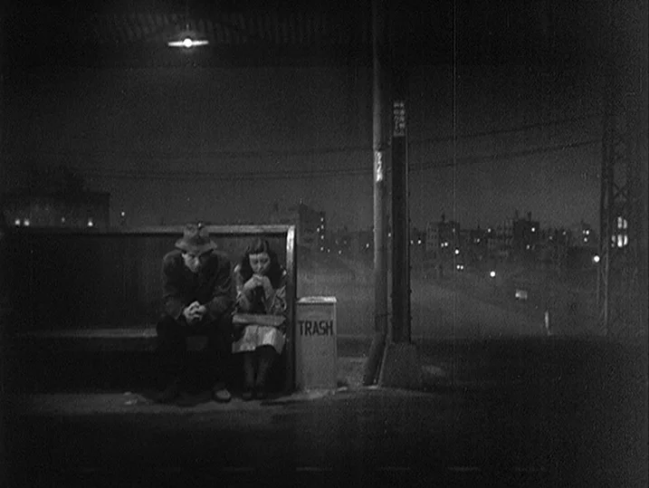 One Wonderful Sunday (1947) movie still: wide — A train station platform at night with a single bench and a trash bin. A man and a woman sit side by…