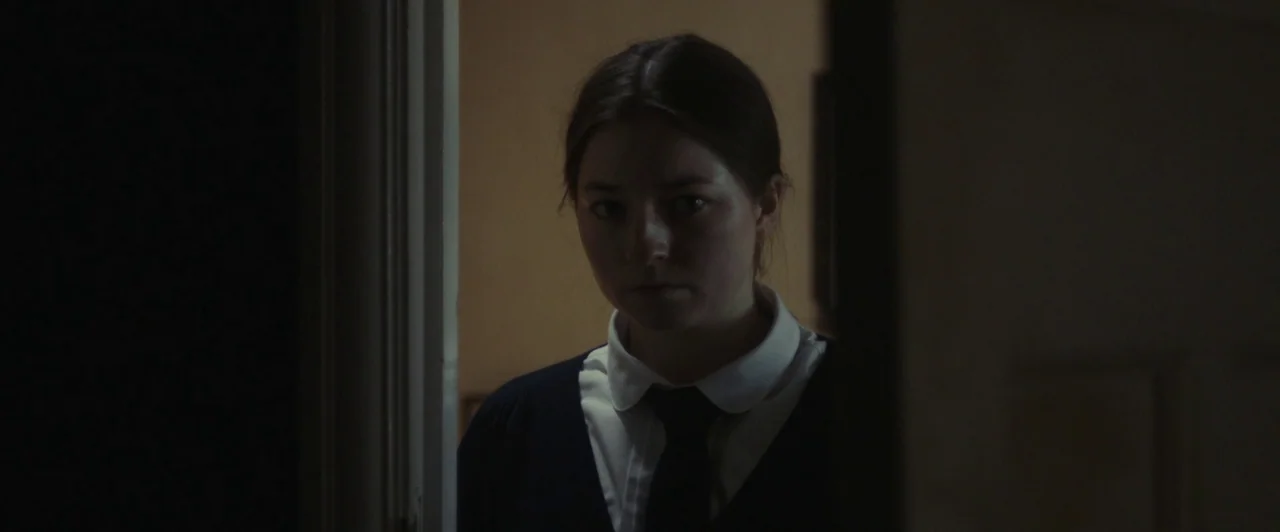 You Are Not My Mother (2021) movie still: medium — A dimly lit room with a person framed within a doorway. A young woman in a school-style uniform…
