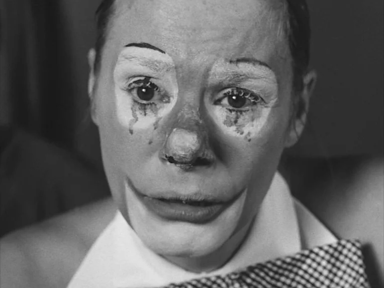The Rite (1969) movie still: closeup — A close-up of a person wearing stage clown makeup in a dimly lit space. The subject stares directly at the…