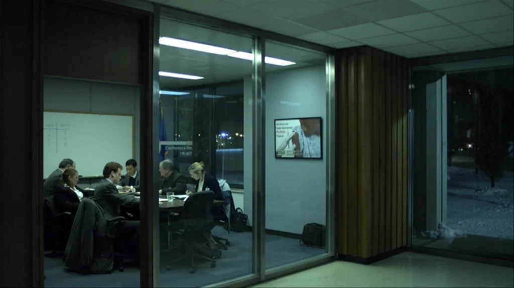 Contagion (2011) movie still: wide — A glass-enclosed conference room features people sitting around a long table at night. A whiteboard stands in…
