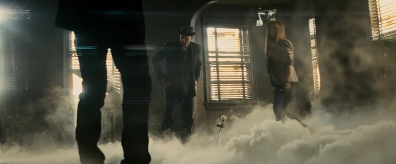 Now You See Me (2013) movie still: wide — A dark, smoke-filled room features two people standing in front of windows with blinds. A single white…