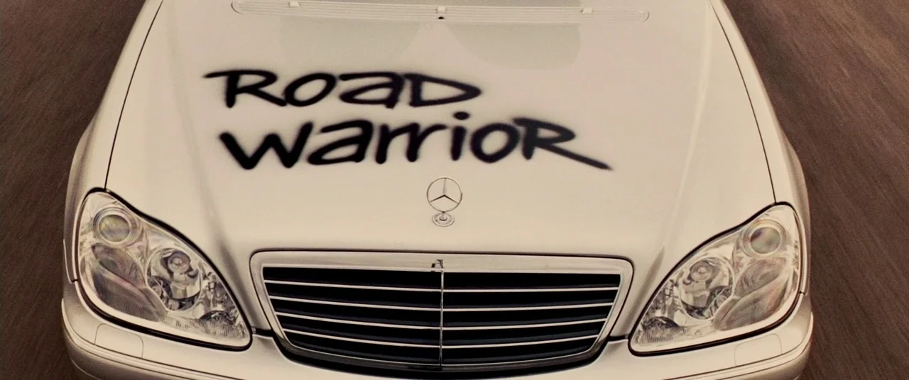 Carriers (2009) movie still: closeup — A silver luxury sedan car hood is marked with spray-painted text in black. The vehicle is captured from a…