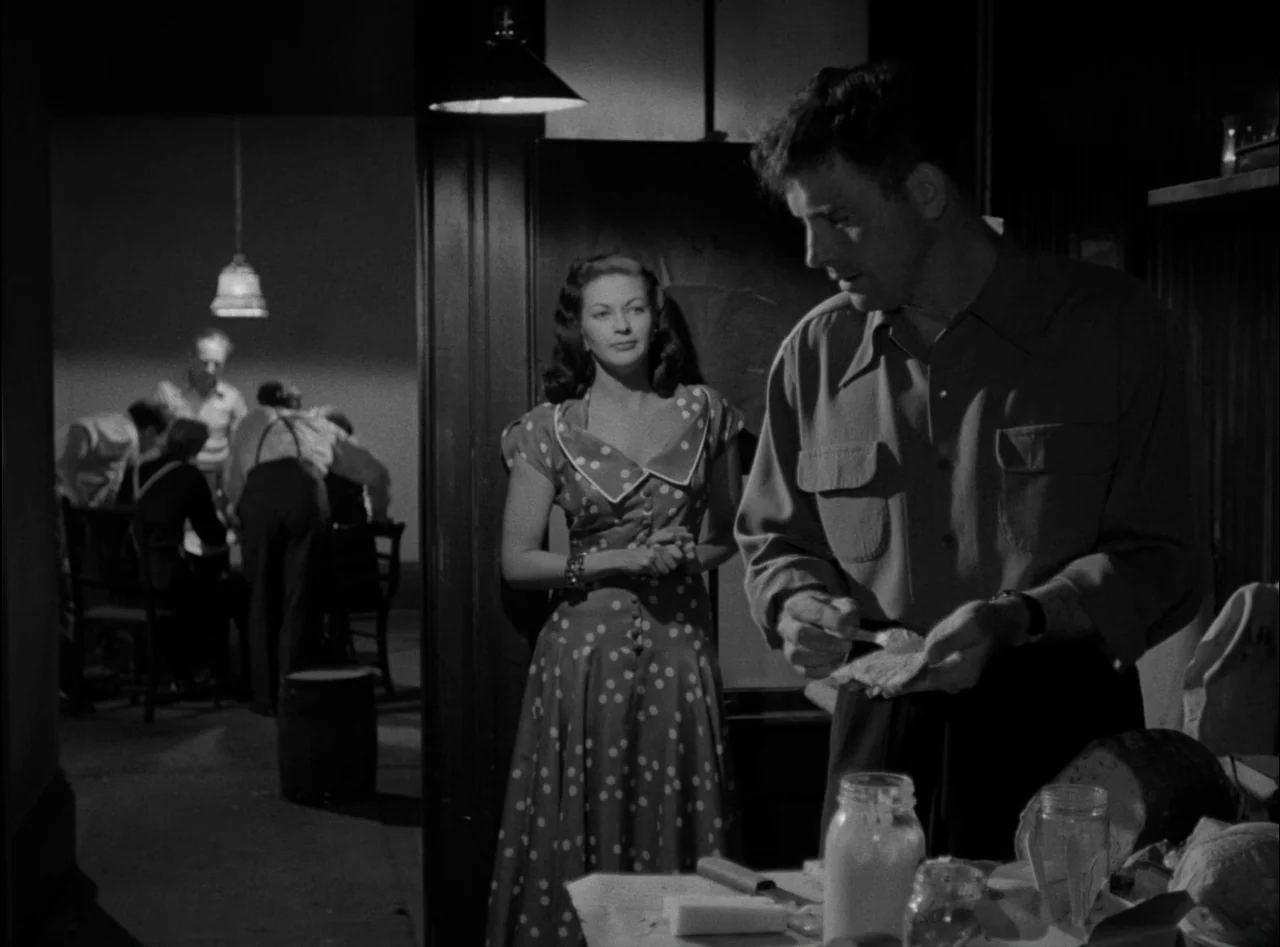 Criss Cross (1949) movie still: medium — A dimly lit room with a man standing at a counter preparing food. A woman stands in the doorway behind him…