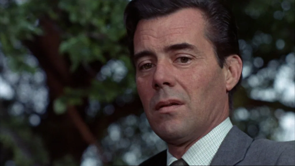 Accident (1967) movie still: closeup — An outdoor setting with lush green foliage blurred significantly in the background. A middle-aged man in a…