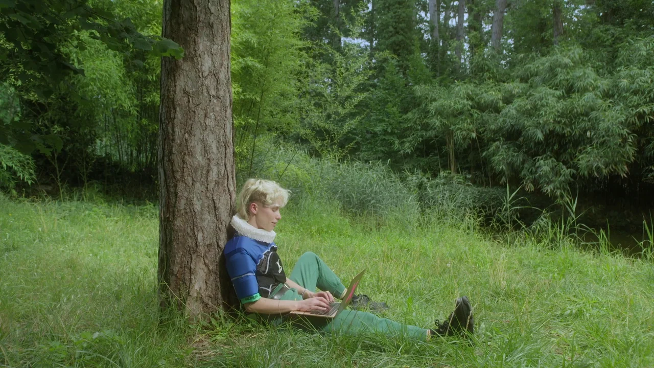 Orlando: My Political Biography (2023) movie still: fullbody — A person sits on the grass at the base of a large tree. They use a laptop computer…
