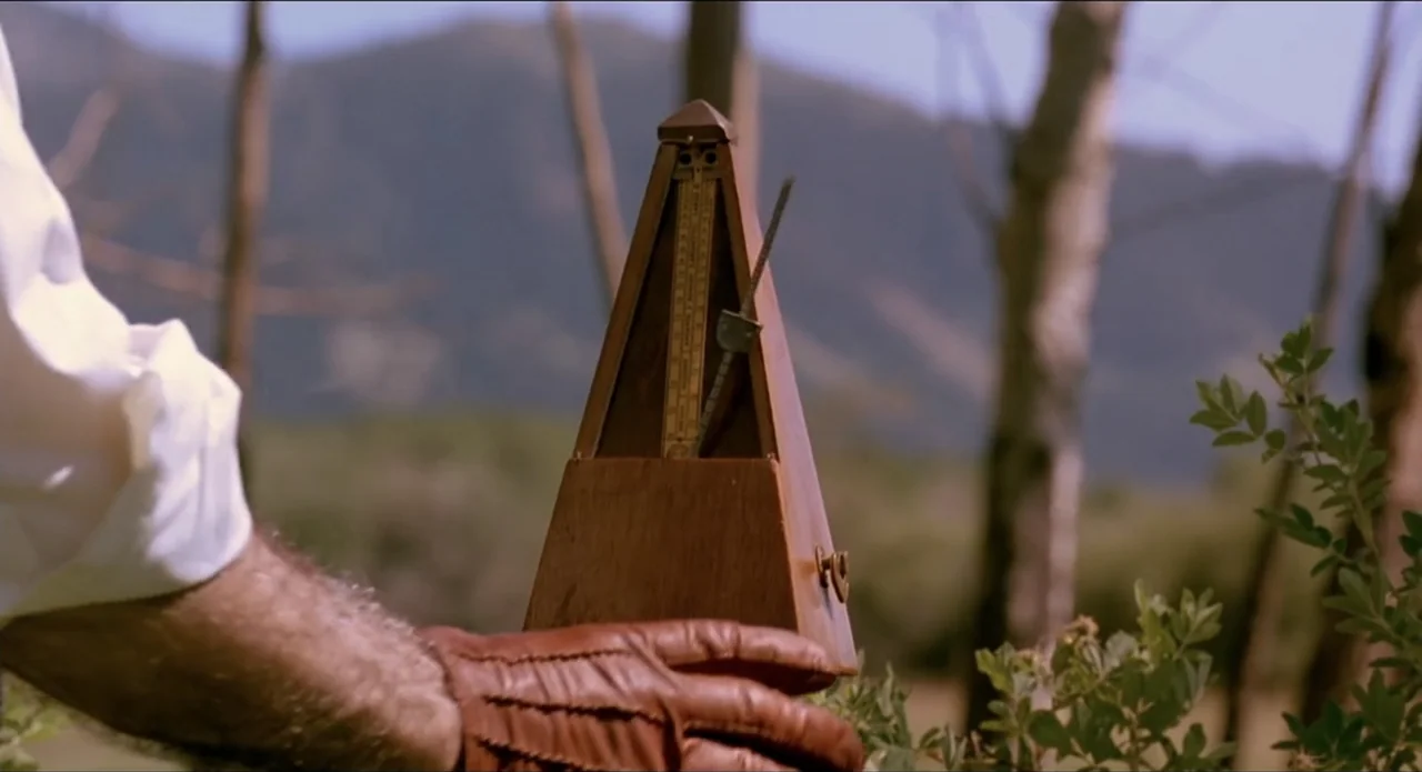 A River Runs Through It (1992) movie still: closeup — A wooden metronome sits in a outdoor field with blurred trees in the background. A man wearing…