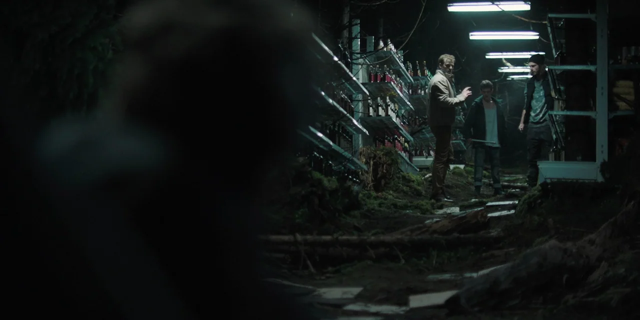 The Ritual (2017) movie still: wide — A surreal outdoor setting where retail store metal shelving units stand among mossy earth. Three men stand…